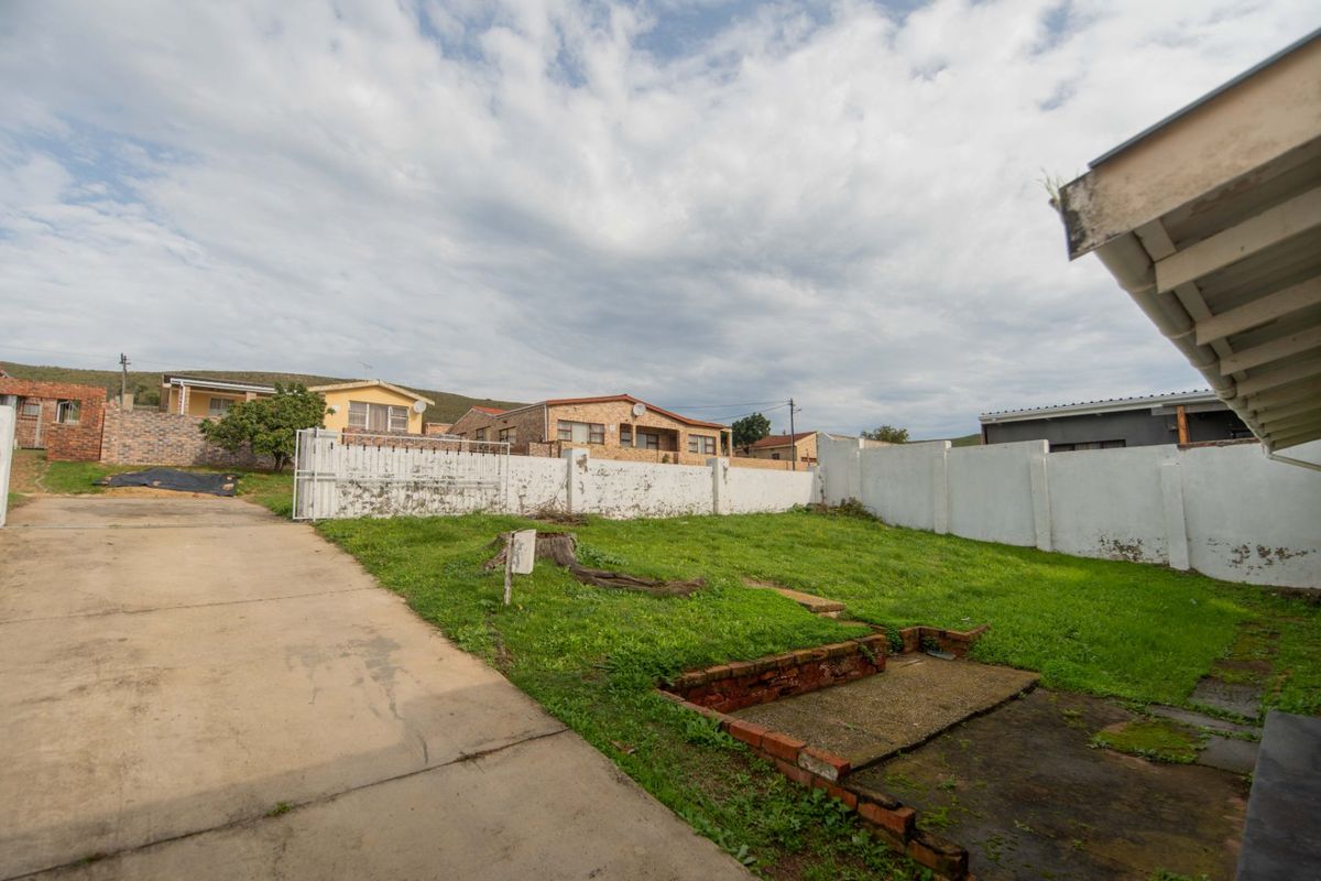 A rear exterior view showing a concrete driveway, a grassed yard area, and a perimeter wall with visible signs of wear.