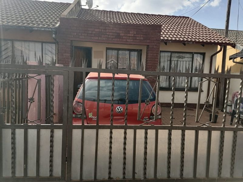 Front elevation of a single-story house featuring a tiled roof, brick facade, and a secure metal gate entrance with a paved driveway area.