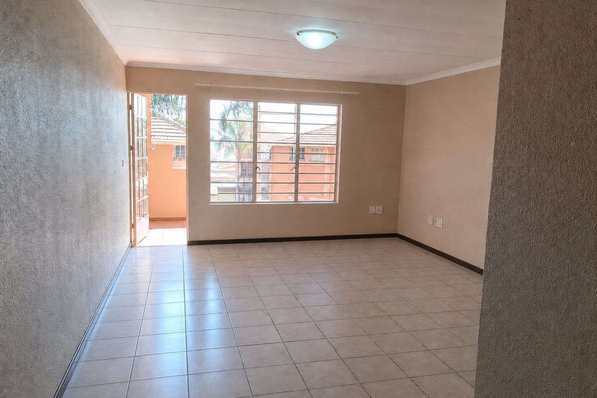 A bright, empty living room featuring neutral tiled flooring, a large window with security bars, and direct access to an outdoor balcony area.