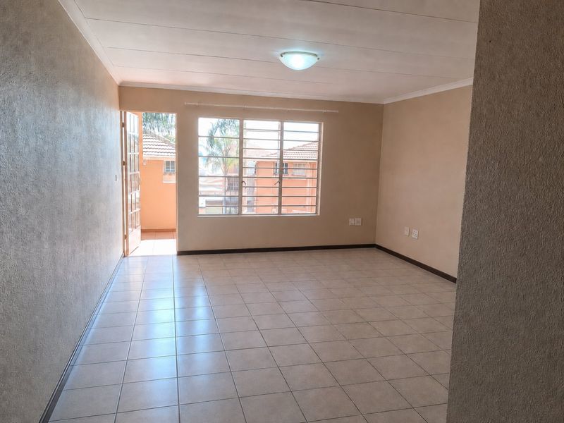 A bright, empty living room featuring neutral tiled flooring, a large window with security bars, and direct access to an outdoor balcony area.