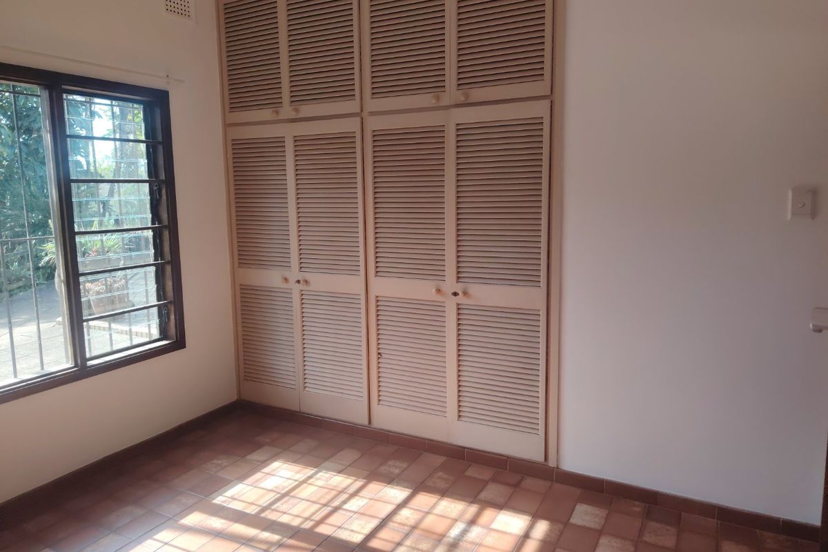 A bright, empty bedroom featuring built-in louvered cupboards, tiled flooring, and a window with security bars.
