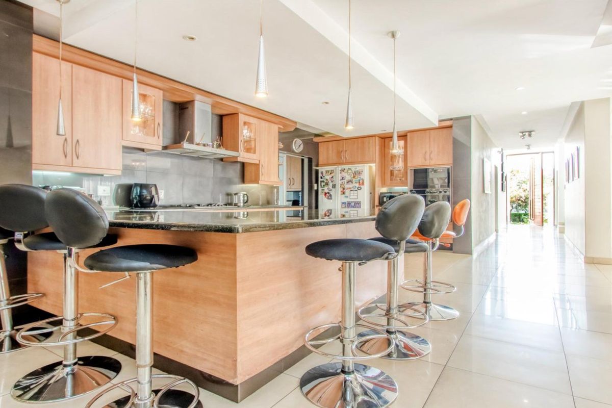 Modern kitchen featuring light wood cabinetry, granite countertops, built-in appliances, and a breakfast bar with pendant lighting, leading into a bright hallway.