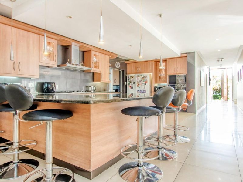 Modern kitchen featuring light wood cabinetry, granite countertops, built-in appliances, and a breakfast bar with pendant lighting, leading into a bright hallway.