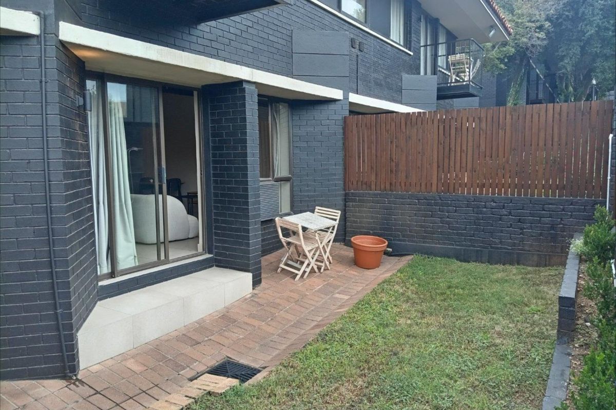 A private rear courtyard featuring a paved patio area, dark brick exterior walls, and a timber privacy fence, accessible via sliding glass doors.