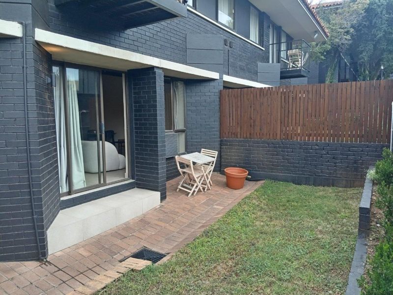 A private rear courtyard featuring a paved patio area, dark brick exterior walls, and a timber privacy fence, accessible via sliding glass doors.