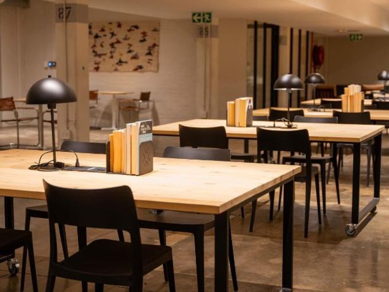 A modern, open-plan communal workspace or clubhouse area featuring polished concrete floors, industrial-style track lighting, and multiple study or meeting tables.