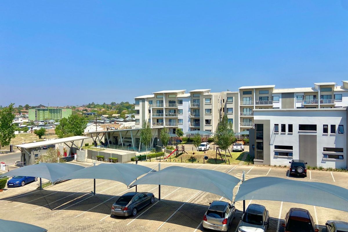 An elevated view of the apartment complex grounds, featuring paved parking with shade-cloth carports, communal play areas, and modern multi-story residential buildings.