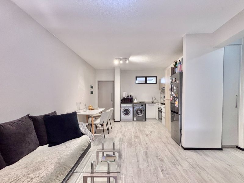 An open-plan living and kitchen area featuring light-toned laminate flooring, modern cabinetry, and integrated kitchen appliances in a functional, well-lit layout.