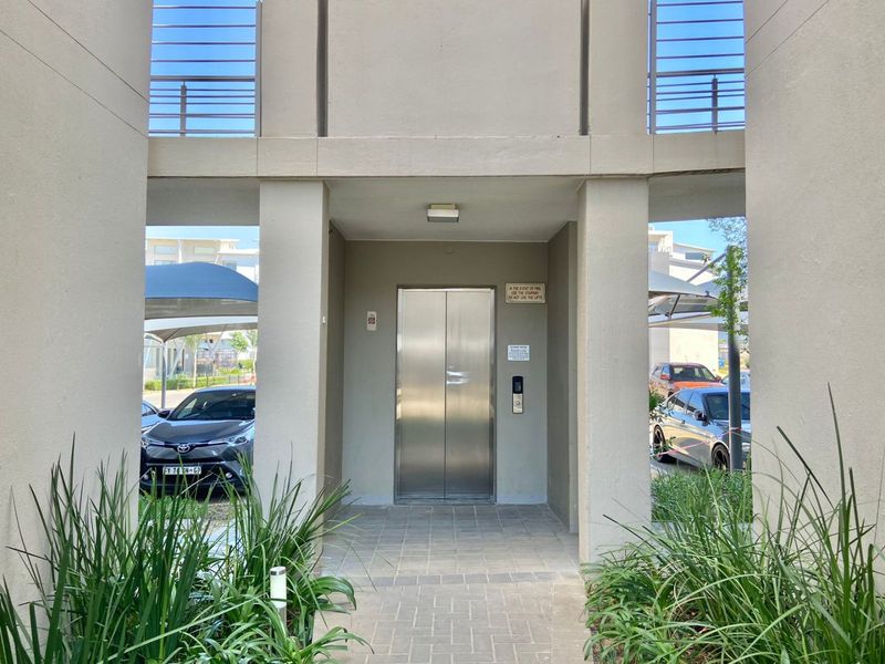 A modern apartment building entrance featuring a stainless steel elevator, paved walkway, and concrete structural pillars.