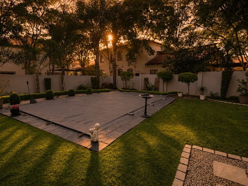 A well-maintained backyard featuring a swimming pool with a safety cover, surrounded by a manicured lawn, landscaped garden beds, and a perimeter wall.