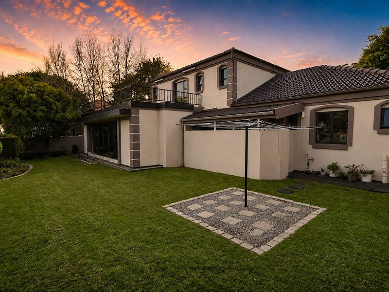 A well-maintained rear exterior view of the house featuring a landscaped garden, paved area, and an upper-level balcony with metal railings at sunset.