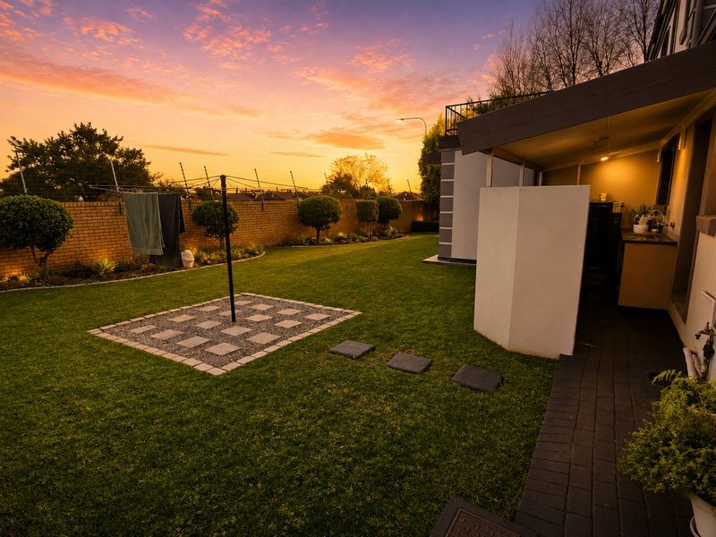 A well-maintained rear garden featuring a paved patio area, manicured lawn, brick boundary wall, and integrated outdoor lighting at sunset.