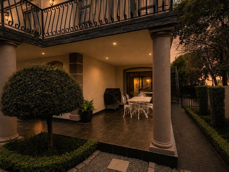 A covered patio area featuring tiled flooring, recessed lighting, and structural columns, with an overhead balcony and access to the garden.