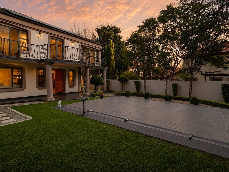 A well-maintained rear exterior view featuring a landscaped garden, a covered swimming pool, and a two-story home with a balcony and classic architectural columns.