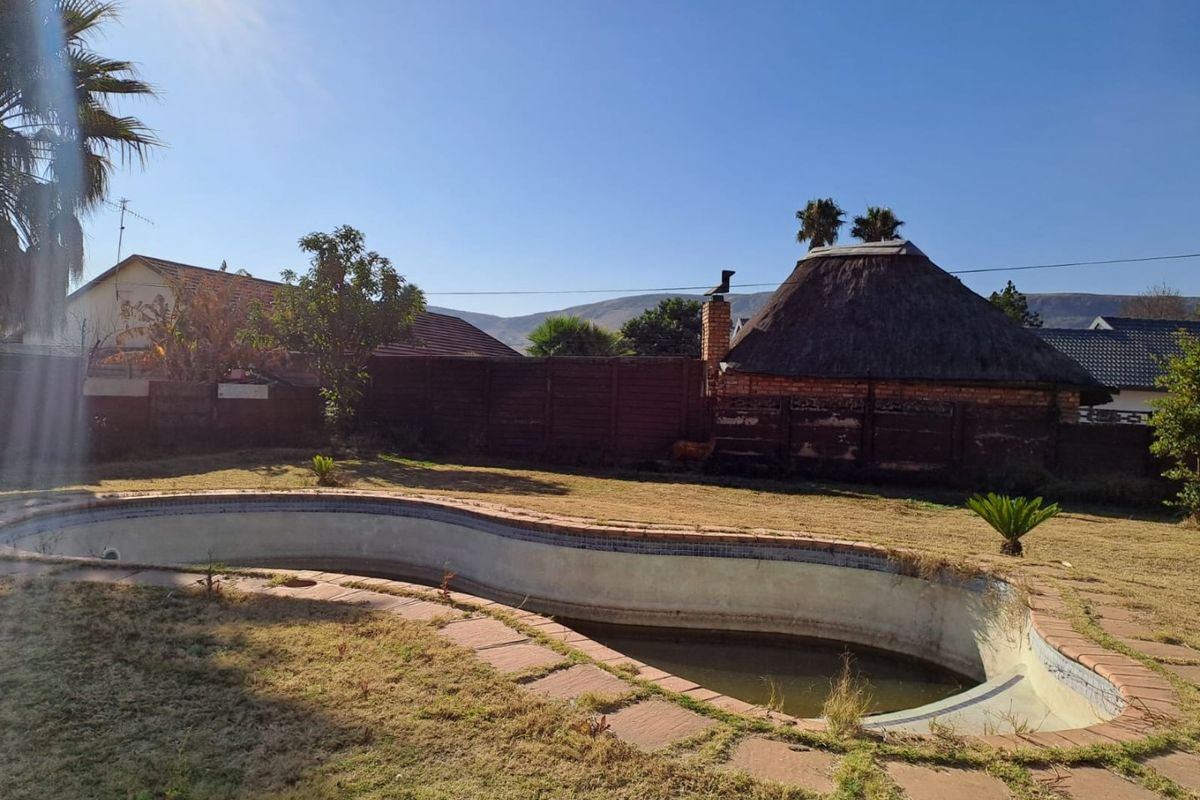 Backyard area featuring an empty swimming pool, paved surround, and a traditional thatched lapa structure set against a boundary wall.