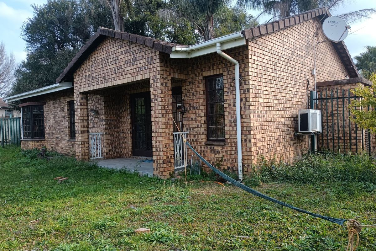 Rear view of a face-brick townhouse featuring a covered patio, security bars on windows, and an external air conditioning unit set in an overgrown garden.