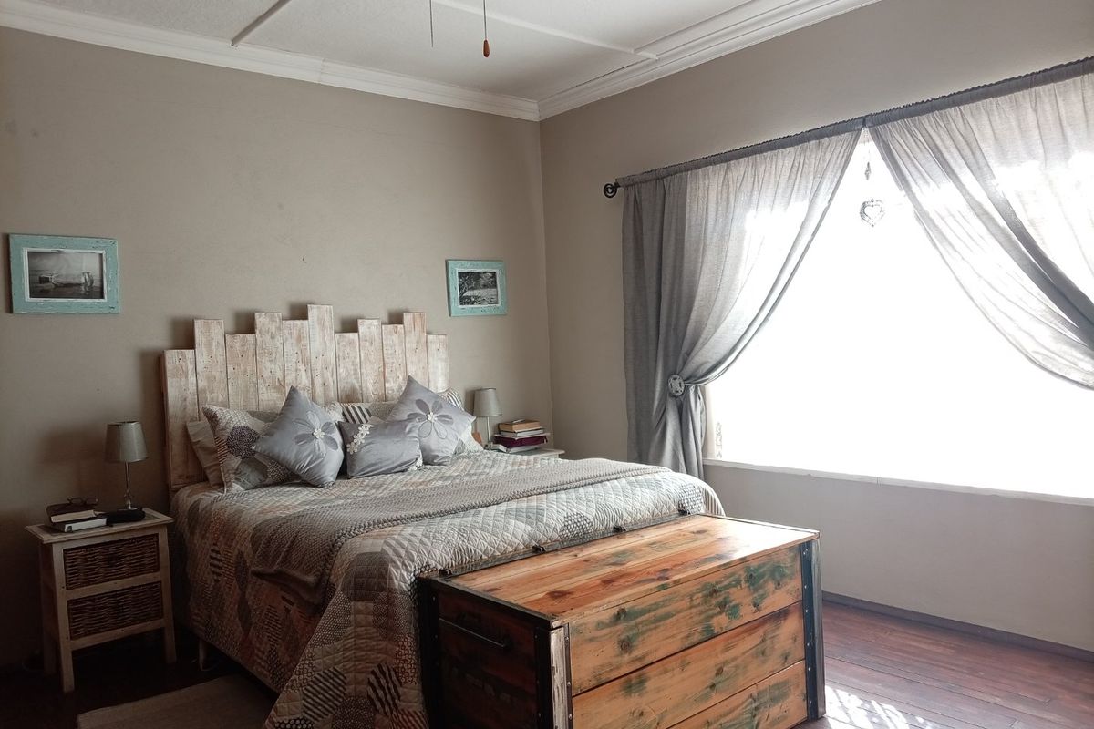 A cozy bedroom featuring timber flooring, crown molding, and a ceiling fan, with natural light entering through a large window.