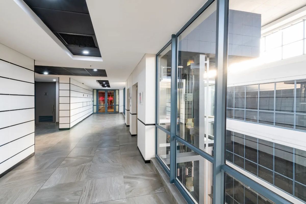 Modern commercial hallway featuring tiled flooring, recessed ceiling lighting, and a prominent glass-enclosed elevator shaft.