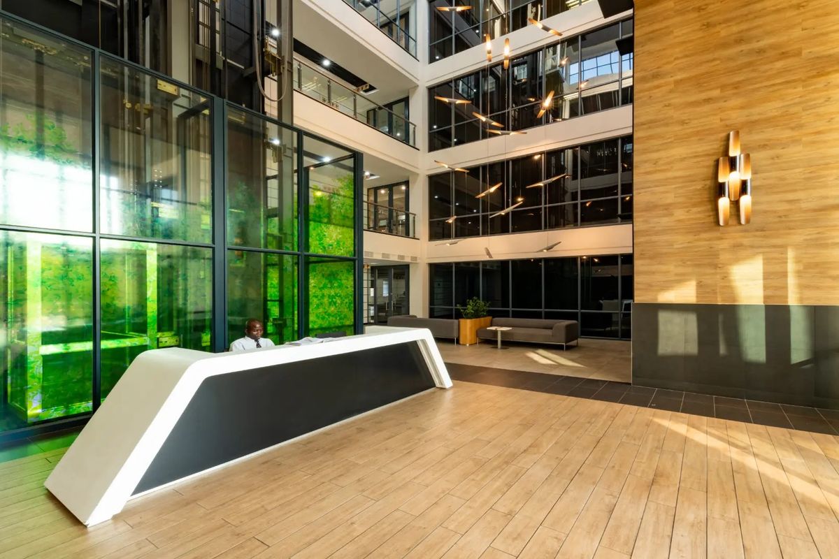 Modern commercial building lobby featuring a high-ceiling atrium, glass elevator shaft, reception desk, and contemporary wood-look finishes.
