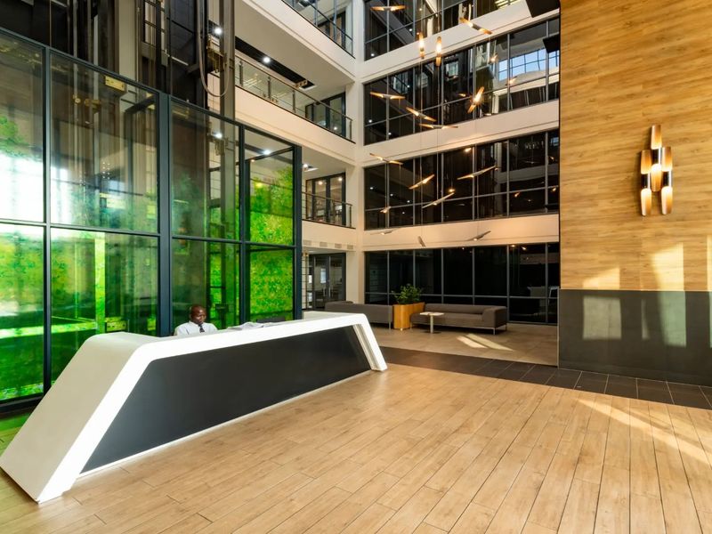 Modern commercial building lobby featuring a high-ceiling atrium, glass elevator shaft, reception desk, and contemporary wood-look finishes.