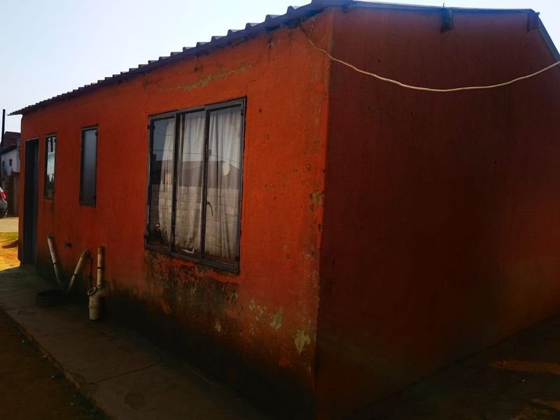 Exterior view of a residential building showing weathered orange painted walls, metal window frames, and external plumbing fixtures.
