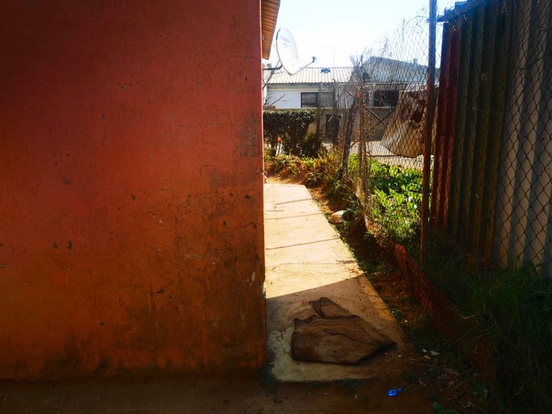 A narrow exterior side passage featuring a concrete walkway, chain-link boundary fencing, and a weathered exterior wall.