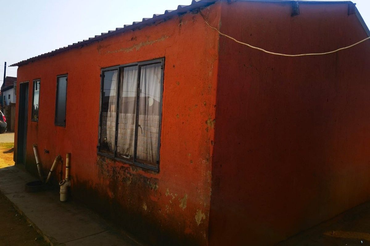 Exterior view of a residential structure showing weathered orange painted walls, metal window frames, and exposed external plumbing.