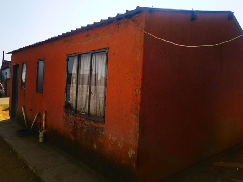 Exterior view of a residential structure showing weathered orange painted walls, metal window frames, and exposed external plumbing.