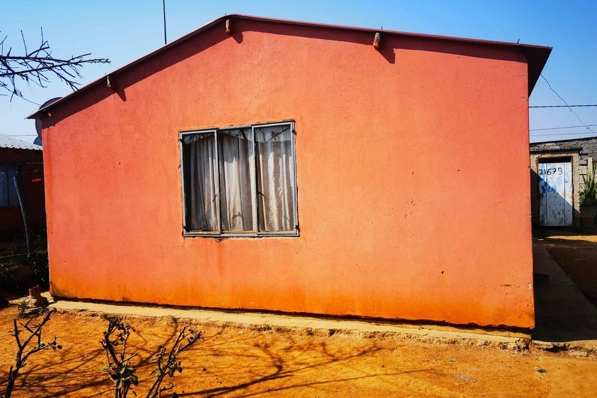 Rear view of a single-story house featuring a simple pitched roof, metal-framed window, and exterior plaster finish on a dirt plot.