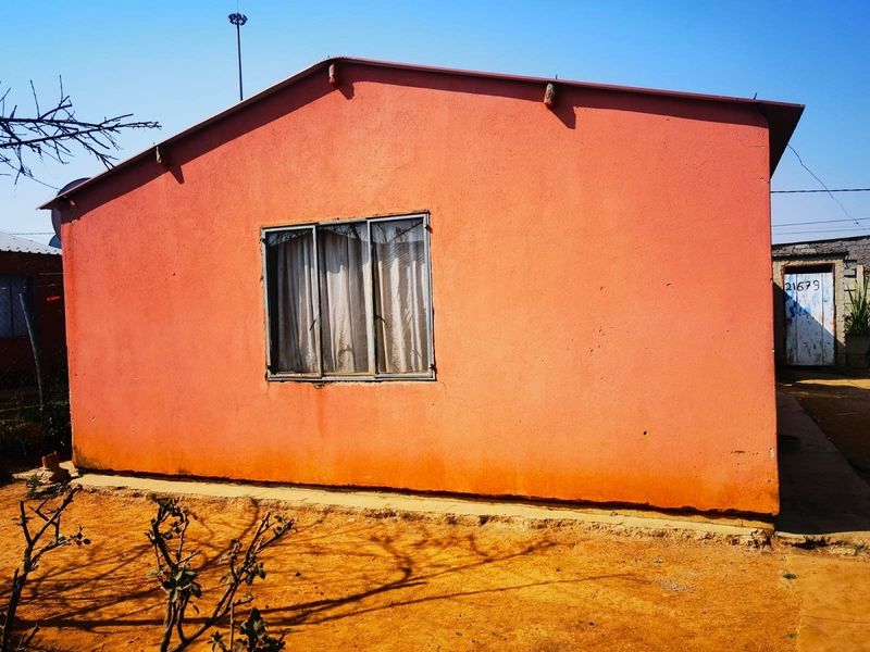 Rear view of a single-story house featuring a simple pitched roof, metal-framed window, and exterior plaster finish on a dirt plot.