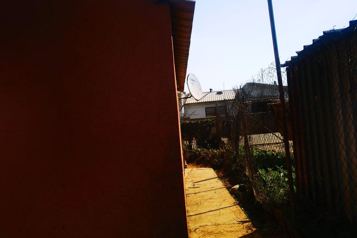 A narrow side passage between the house exterior and a chain-link boundary fence, featuring a concrete path and mounted satellite dish.