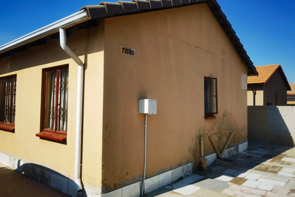 Rear exterior view of a single-story house featuring tiled roofing, security bars on windows, and a paved patio area with visible wall maintenance requirements.