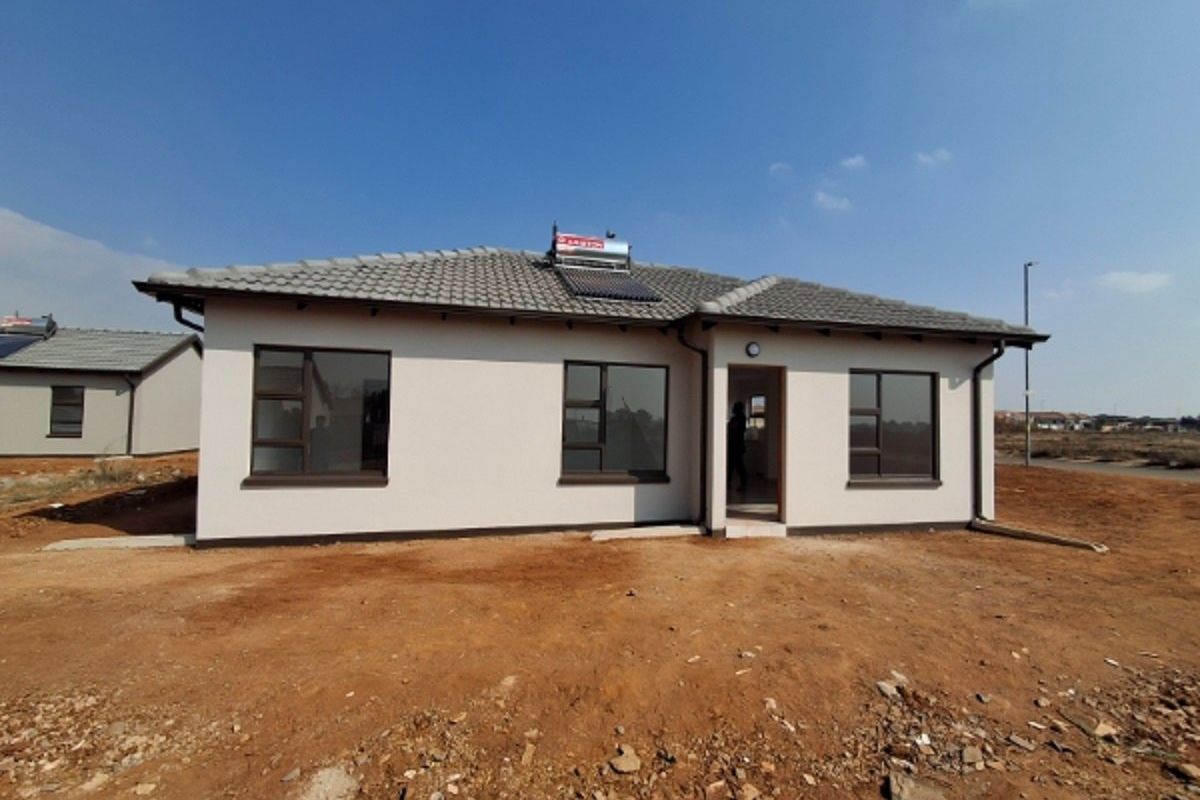 A modern single-story residential house featuring a tiled roof, solar water heating system, and aluminium window frames, set on an unlandscaped plot.