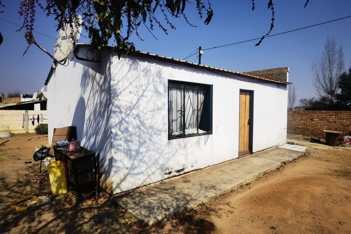 Rear exterior view of a small residential structure featuring a concrete walkway, metal security window bars, and a corrugated metal roof.
