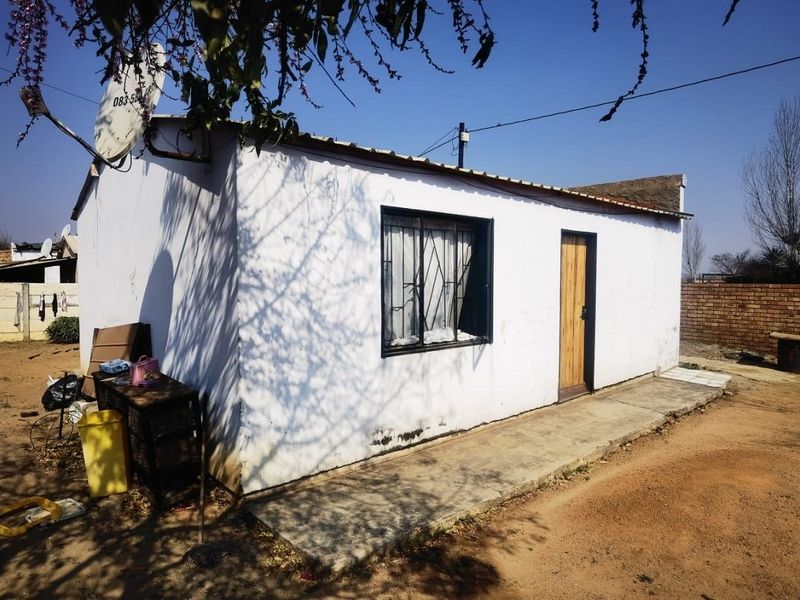 Rear exterior view of a small residential structure featuring a concrete walkway, metal security window bars, and a corrugated metal roof.