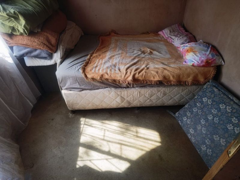 A compact bedroom featuring unfinished wall surfaces and exposed concrete flooring, with natural light entering through a window with security bars.