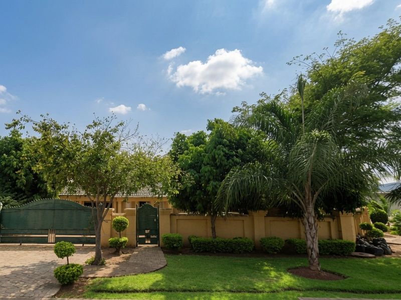 Front view of the property featuring a secure perimeter wall, gated driveway entrance, paved walkway, and well-maintained landscaped garden.