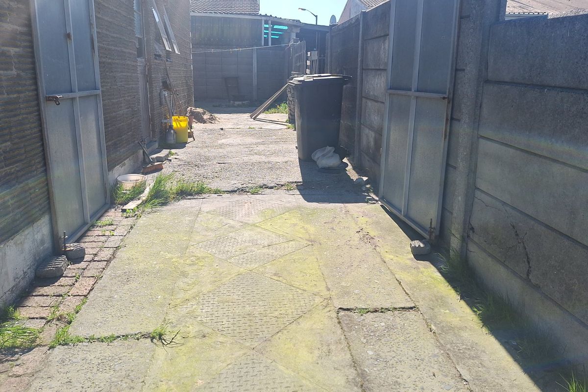 A narrow rear exterior area featuring concrete paving, metal gates, and precast boundary walls, showing signs of wear and weathering.