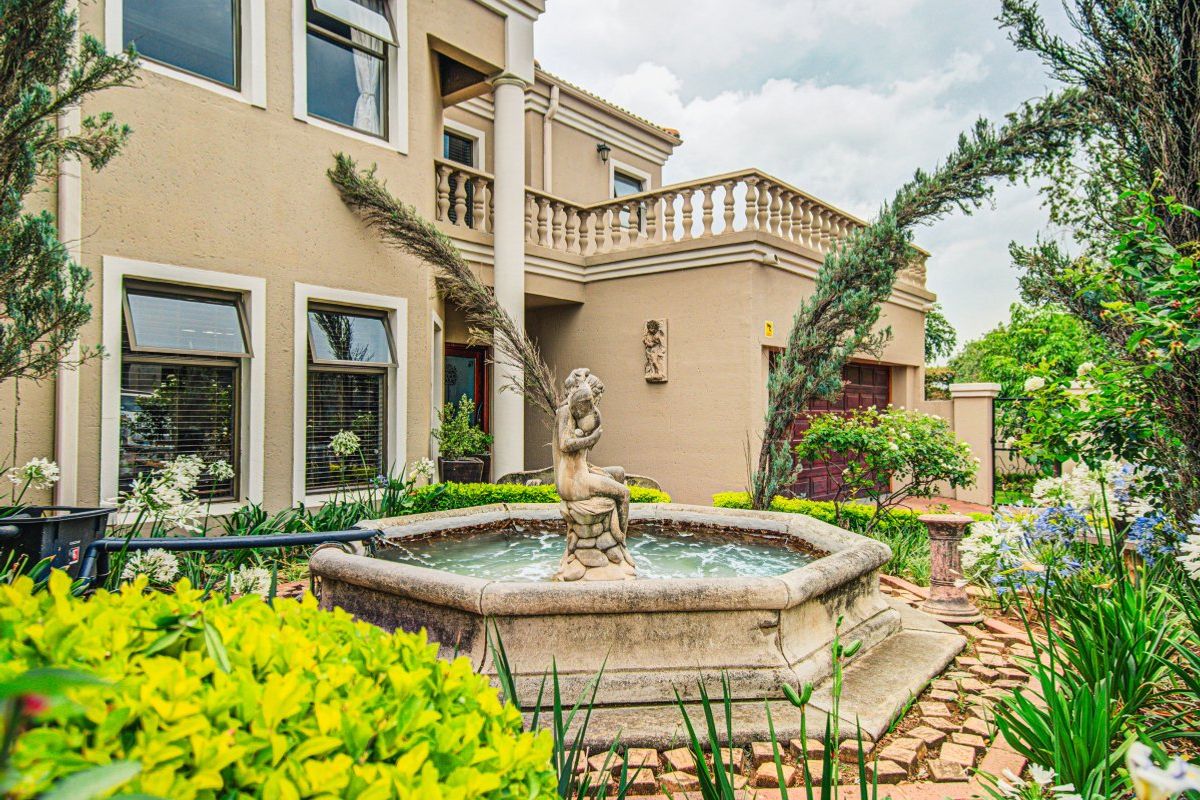 A well-maintained rear garden featuring a stone fountain, paved pathways, and a classic architectural facade with a balcony and double garage.