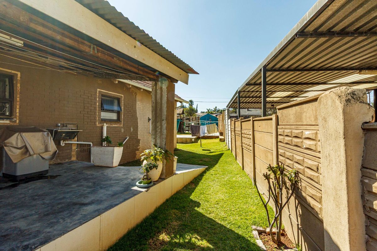Rear exterior view featuring a covered patio area, concrete boundary wall, and a side carport structure with a lawn.
