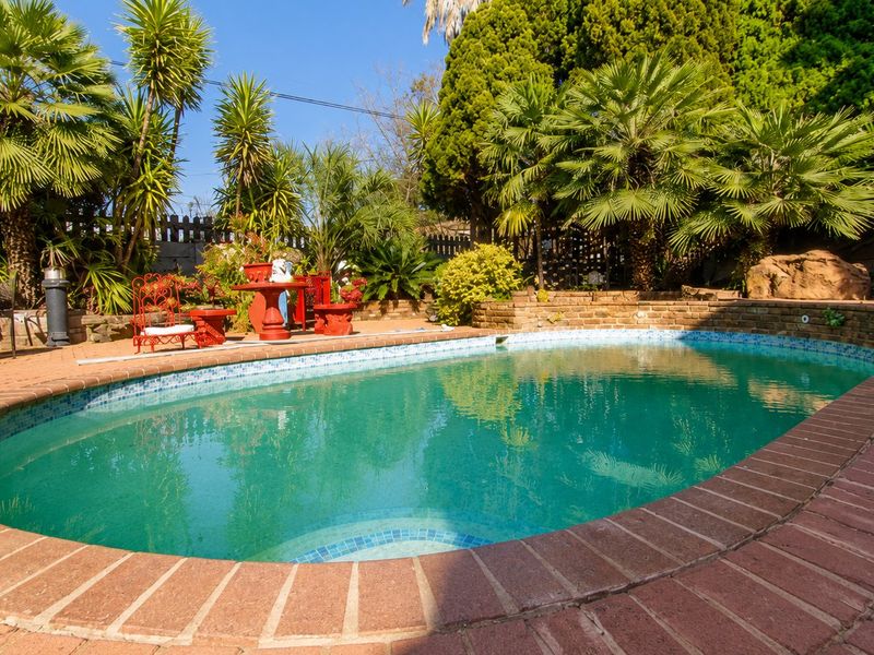 A well-maintained outdoor swimming pool area featuring brick coping, a surrounding brick retaining wall, and established tropical landscaping.