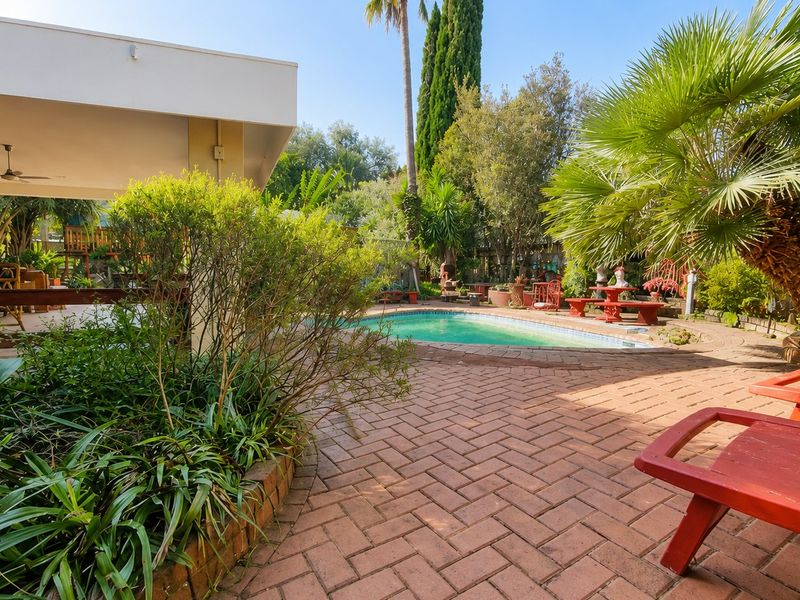 Outdoor entertainment area featuring a swimming pool, brick-paved patio, and a covered veranda with a ceiling fan, set within a mature garden.