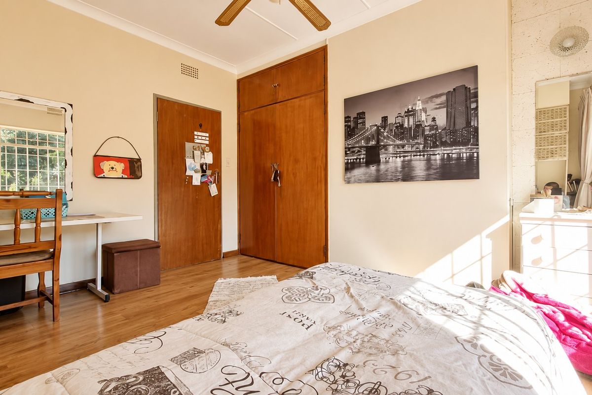 A bedroom featuring laminate flooring, built-in wooden cupboards, and a ceiling fan, offering a functional space with natural light.