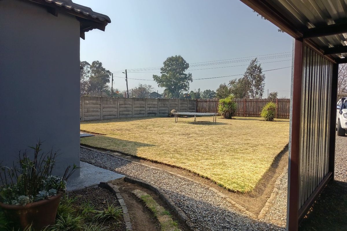 A spacious rear garden featuring a lawn, gravel pathways, and a precast concrete boundary wall, viewed from under a metal carport structure.