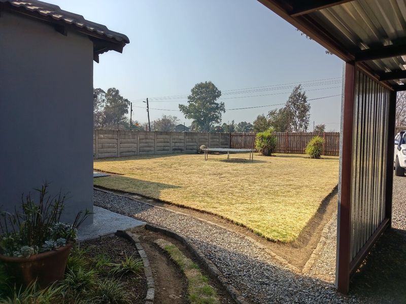 A spacious rear garden featuring a lawn, gravel pathways, and a precast concrete boundary wall, viewed from under a metal carport structure.