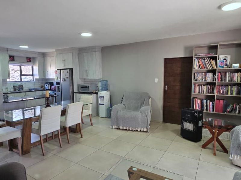 Open-plan living and dining area featuring tiled floors, recessed lighting, and a large built-in shelving unit, seamlessly connected to a functional kitchen space.