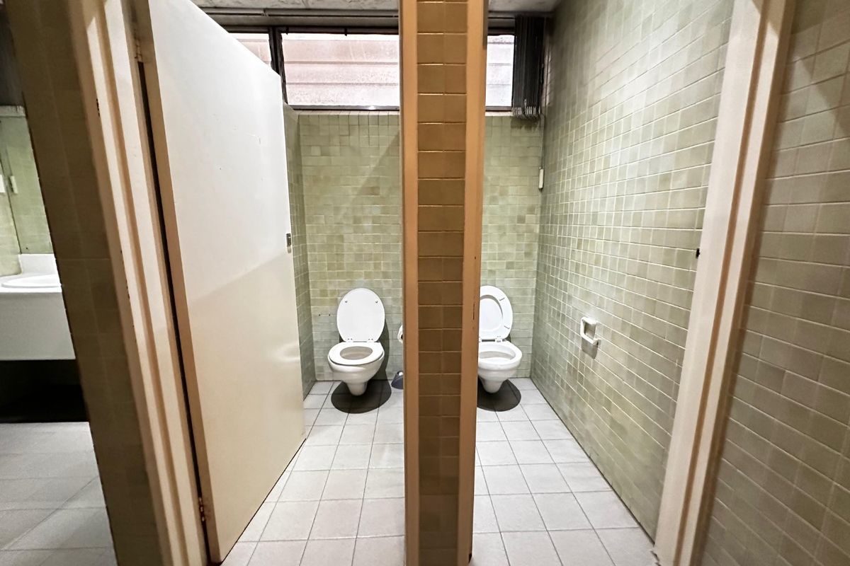 Commercial bathroom facility featuring two toilet stalls with tiled walls and flooring, requiring modernization to improve the overall aesthetic and condition.