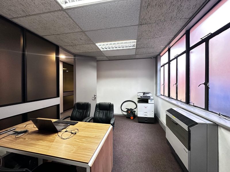 A functional commercial office space featuring a suspended ceiling, recessed lighting, large windows for natural light, and a wall-mounted HVAC unit.