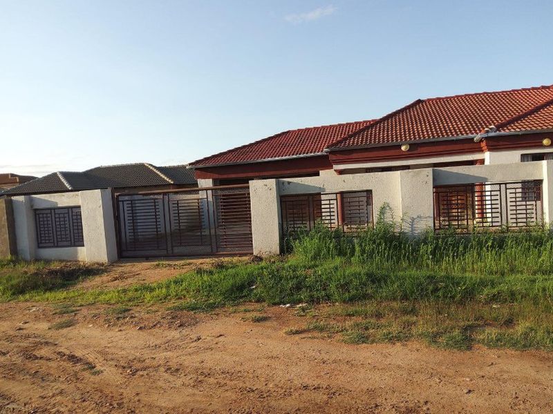 Front exterior view of a single-story house featuring a tiled roof, perimeter wall, and metal security gates, situated on an unpaved road.