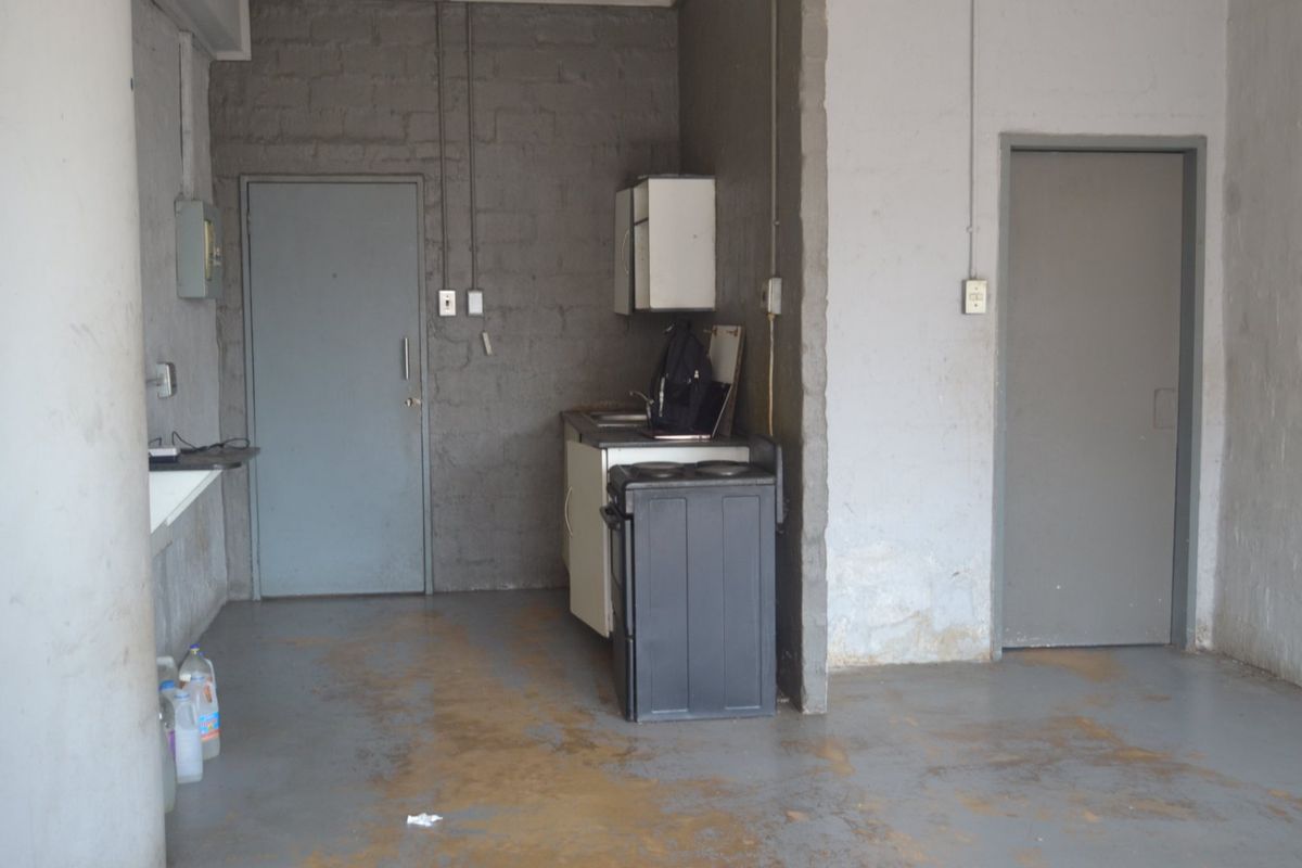 A basic, utilitarian kitchen area featuring exposed brick walls, a small sink unit, a stove, and painted concrete flooring in need of refurbishment.